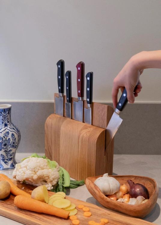 En hand tar en kniv ur ett knivblock i trä på en köksbänk med blomkål, morötter, lök och vitlök.