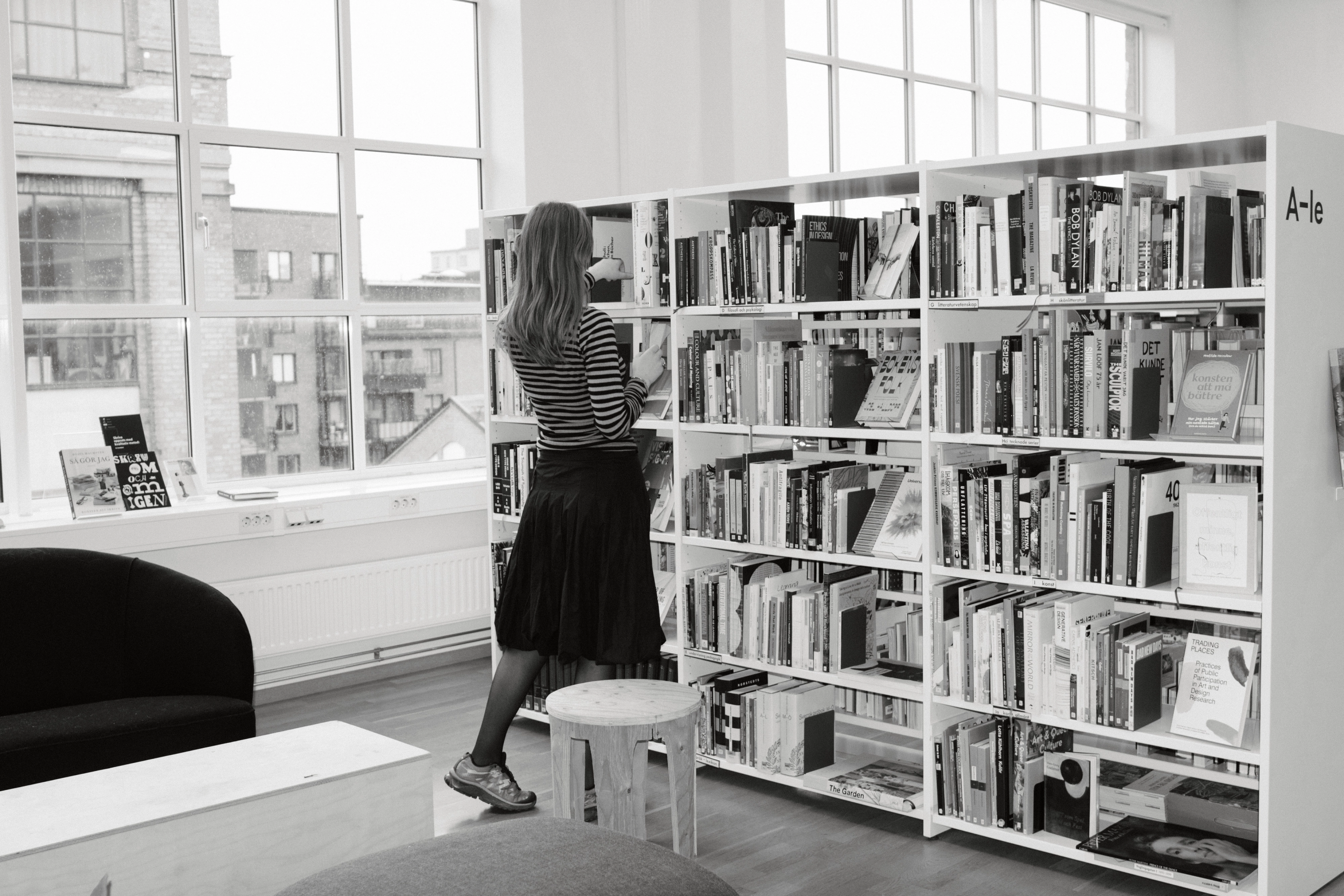Svartvitt: person i randig tröja letar bland höga vita bokhyllor i ett bibliotek med stora fönster.