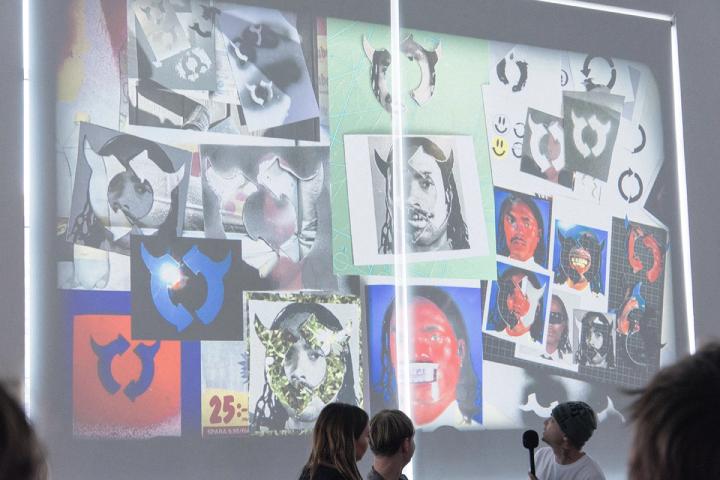 Person med mikrofon presenterar framför en projektion av ett collage med manipulerade porträtt och återkommande pilsymboler.
