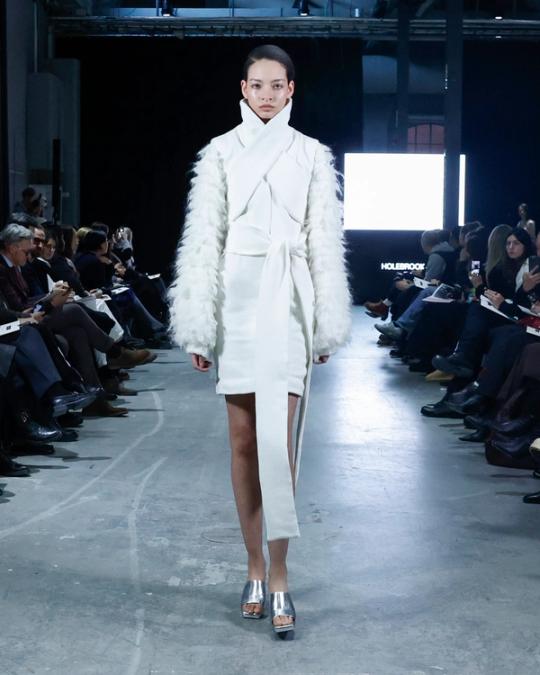 Modell på catwalk i vit omlottkappa med hög krage och fluffiga ärmar, och silverfärgade skor; publik på båda sidor.