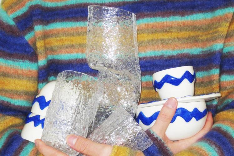 Person i randig tröja håller strukturerade klara glaskärl och vita koppar med blå sicksackränder mot bröstet.