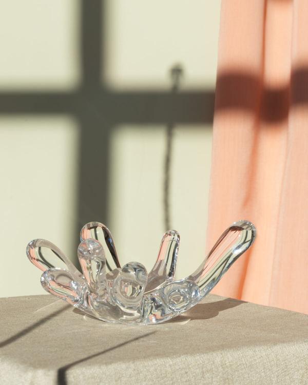 Glasföremål i klart glas med utåtböjda armar, likt en öppen hand eller blomma, på ett podium bredvid en rosa gardin, fönsterskuggor på väggen.
