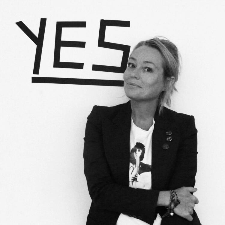 Svartvitt porträtt av en kvinna i kavaj och tryckt t-shirt, med ordet "YES" i stora bokstäver på väggen bakom.