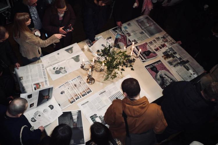 Besökare samlas kring ett bord fullt av tidningar gjorda som studentprojekt, med tända ljus och grönska i mitten, sett ovanifrån.