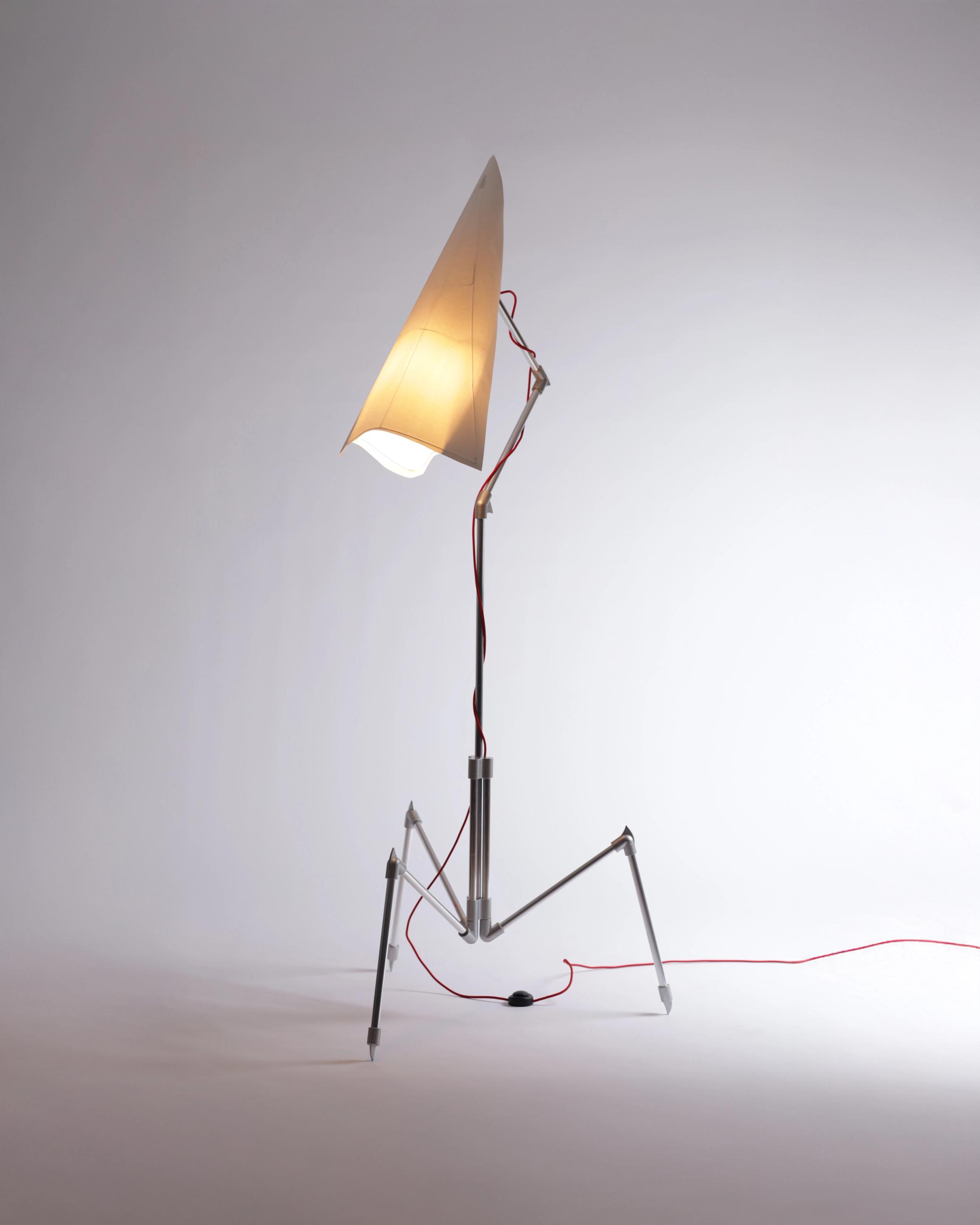 Tänd golvlampa inspirerad av barndomsteckningar av utomjordingar och robotar, med konformad pappersskärm på smalt trebent metallstativ.