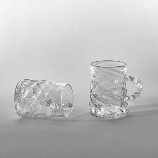 Två vridna glas i klart glas, det ena liggande och det andra stående med vriden handtag, mot ljusgrå bakgrund.