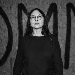 Svartvitt porträtt av en kvinna med långt mörkt hår och runda glasögon i svart tröja, framför en vägg med stora målade bokstäver.