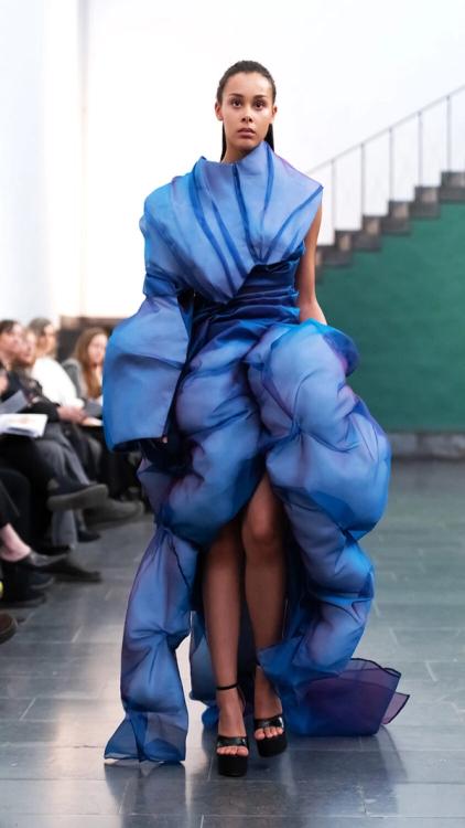 Modell på catwalk i en skulptural blå klänning av veckade, skimrande organzarullar och svarta klackar.Model on a runway in a sculptural blue dress of gathered, iridescent organza rolls and black heels.