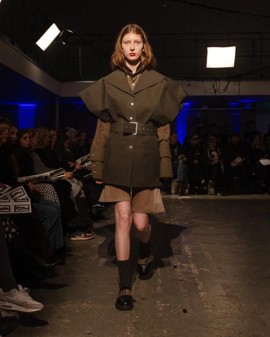 Modell på catwalk i olivgrön kavaj med breda axlar och bälte över beige shorts, svarta strumpor och skor.