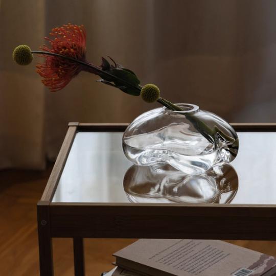 Oregelbundet formad glasvas med en orange nålkuddsprotea och gula bollblommor, på ett glasbord med en bok under.