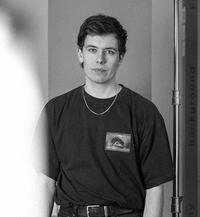 Svartvitt porträtt av en man i mörk t-shirt med ett tryckt öga på bröstet och en halskedja, bredvid ett ljusstativ.