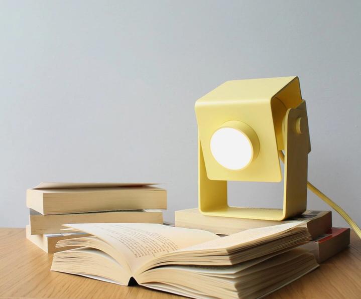 Ljusgul kubformad skrivbordslampa med vinklingsbart huvud, tänd och placerad på en bokhög bredvid en uppslagen bok.