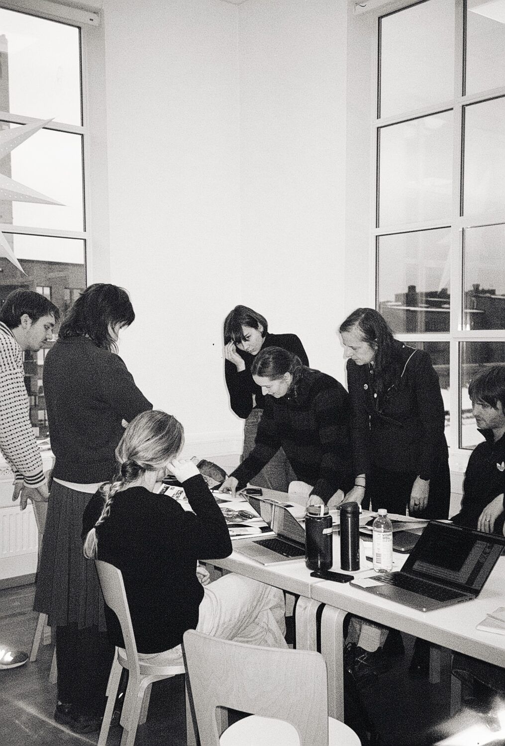 Balch and white: A group of seven people stands around a table, looking at whats laying there. Big window behind.