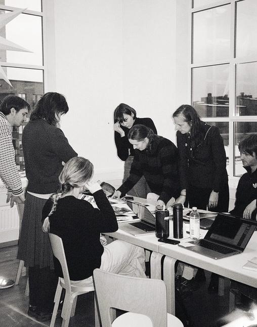 Balch and white: A group of seven people stands around a table, looking at whats laying there. Big window behind.