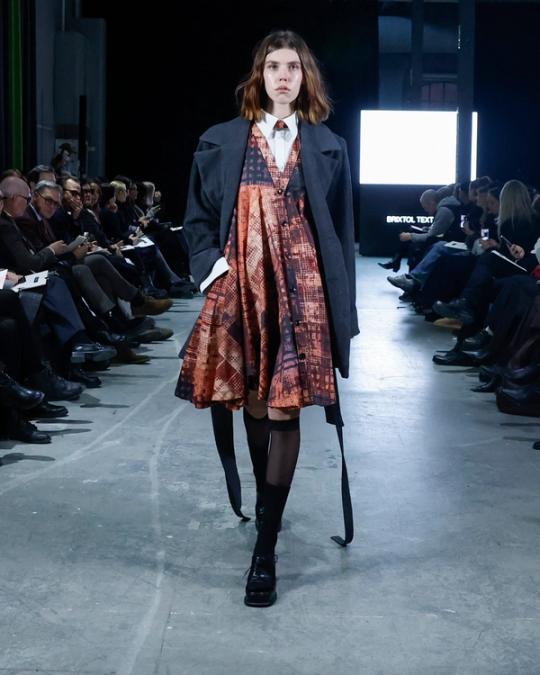 Modell på catwalk i mörk kavaj över en mönstrad klänning i rost och blått, vit skjorta med slips och svarta skor; publik på båda sidor.
