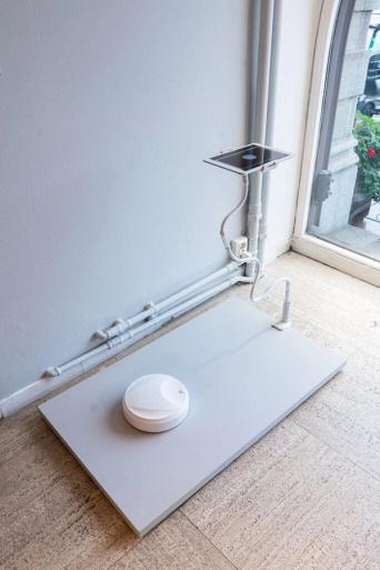 Vit rund robot på en låg grå plattform, kopplad med slangar och rör till en liten skärm på väggen, vid ett fönster.