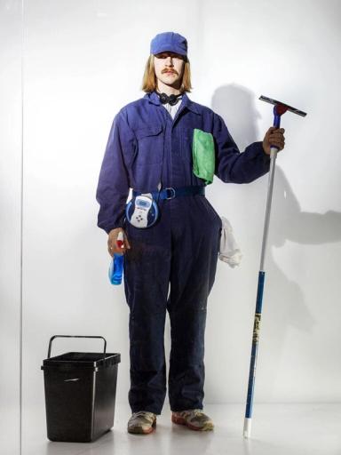Modell utklädd till fönsterputsare i blå overall och keps, med fönsterskrapa, sprayflaska, trasa och en hink bredvid.