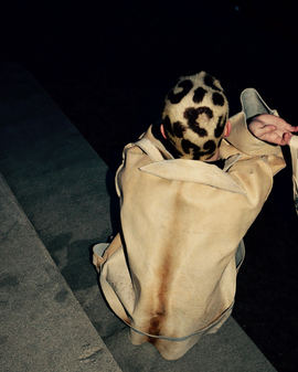 Person med leopardmönstrat rakat huvud sitter med ryggen mot kameran i en beige skinnjacka, på en trappa i mörker. Person with a leopard-patterned shaved head sits with their back to the camera in a beige leather jacket, on steps in the dark.