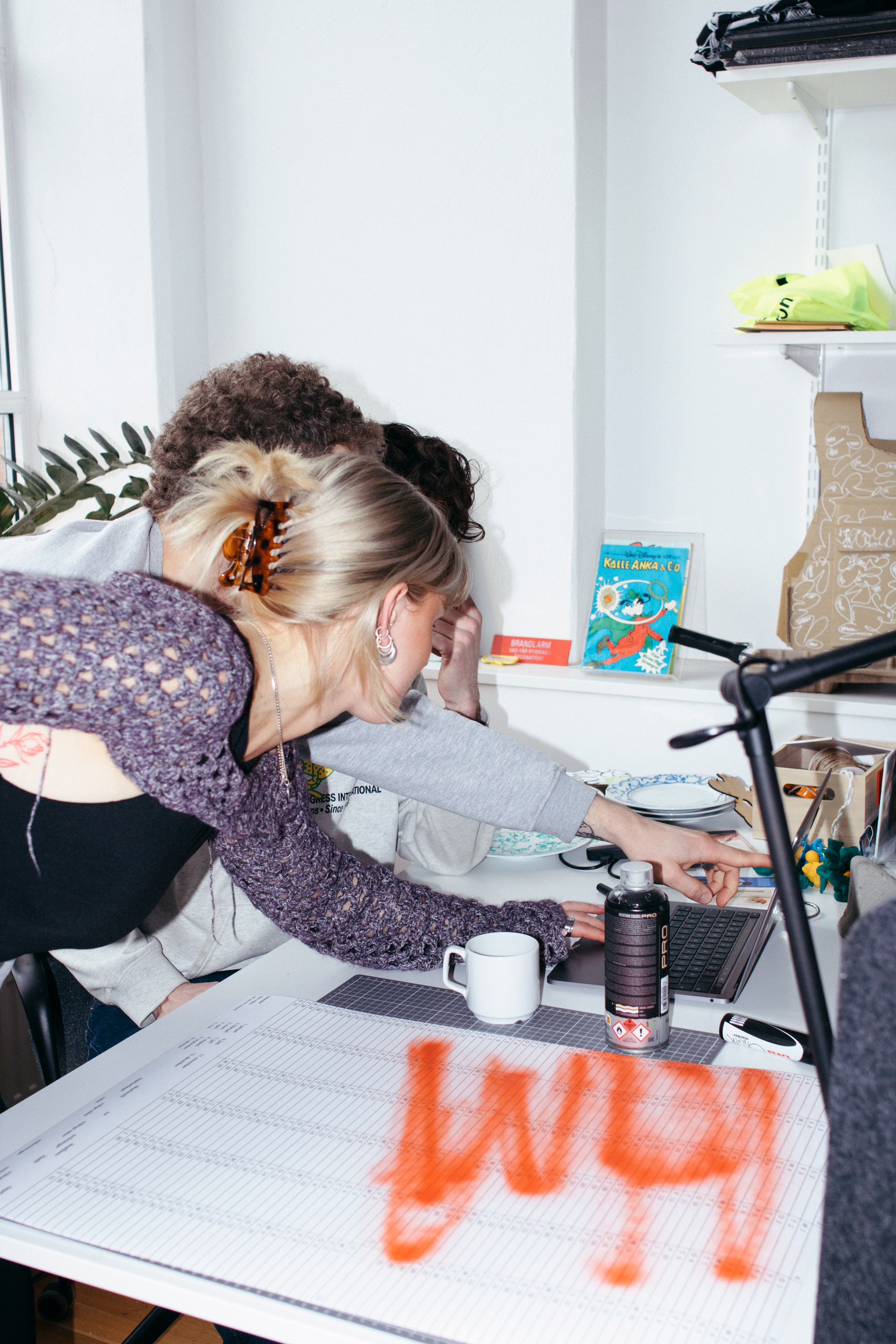 Två personer lutar sig tätt över en laptop vid ett skrivbord, med en sprayburk och ett ark med orange sprejfärg i förgrunden.