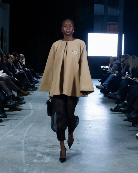 Modell på catwalk i vid kamelfärgad cape, genomskinliga svarta byxor och svarta klackar; publik på båda sidor.