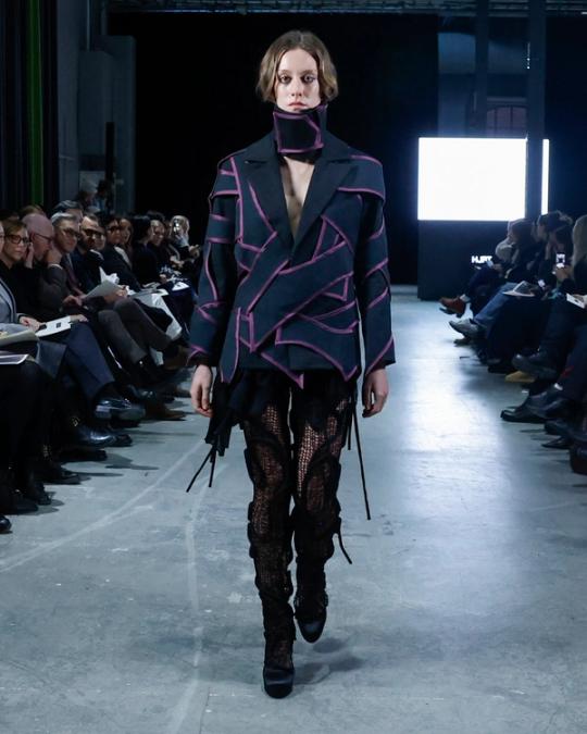 Modell på catwalk i mörkblå kavaj med rosa kantband i vinklade linjer, hög krage, svarta nätbyxor och stövlar; publik på båda sidor.