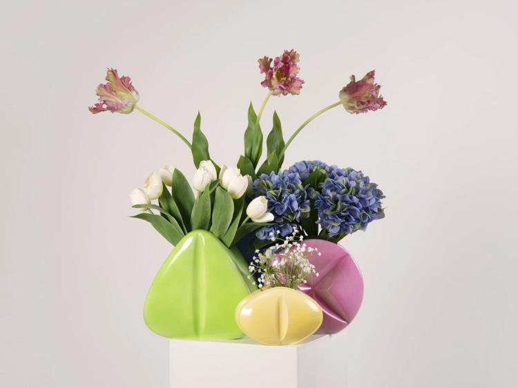 Tre rundade vaser i grönt, gult och rosa med tulpaner, papegojtulpaner, hortensior och slöjblomster, på ett vitt podium.