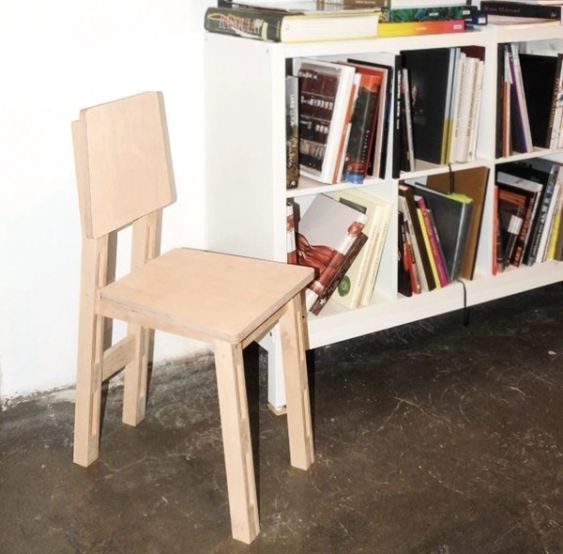 Avskalad stol i ljus plywood, på ett betonggolv bredvid en vit bokhylla full med böcker.