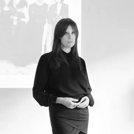 Svartvitt porträtt av en kvinna med långt mörkt hår i svart blus och kjol, framför ett stort fotografi på väggen.