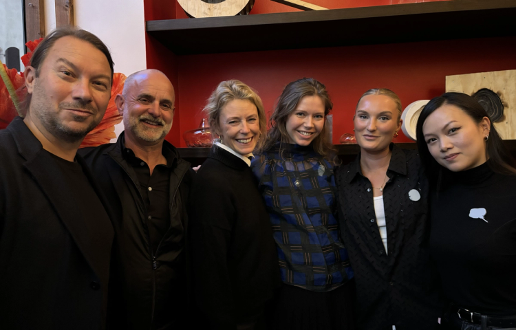 Sex leende personer i mörka kläder står tätt ihop för ett gruppfoto framför en röd vägg med hyllor.