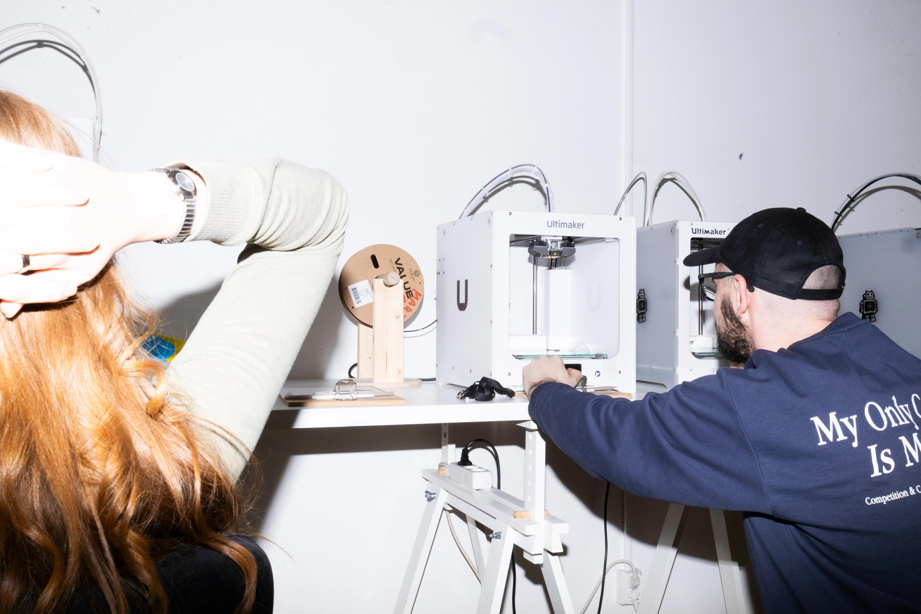 Person i keps justerar en 3D-skrivare bland flera på en hylla medan en annan tittar på, med händerna bakom huvudet.