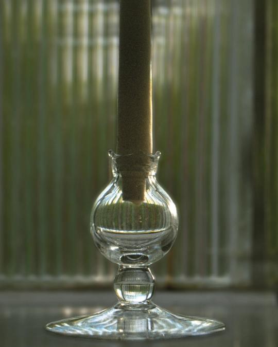 Ljusstake i klart glas med en rund kupa och ett vitt ljus, framför räfflat glas.