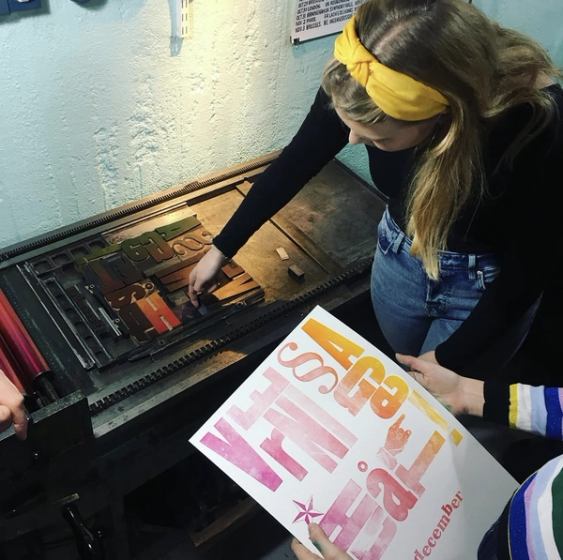 Person placerar trätyper i en boktryckarpress medan ett färgglatt tryck i rosa och gult hålls upp bredvid.