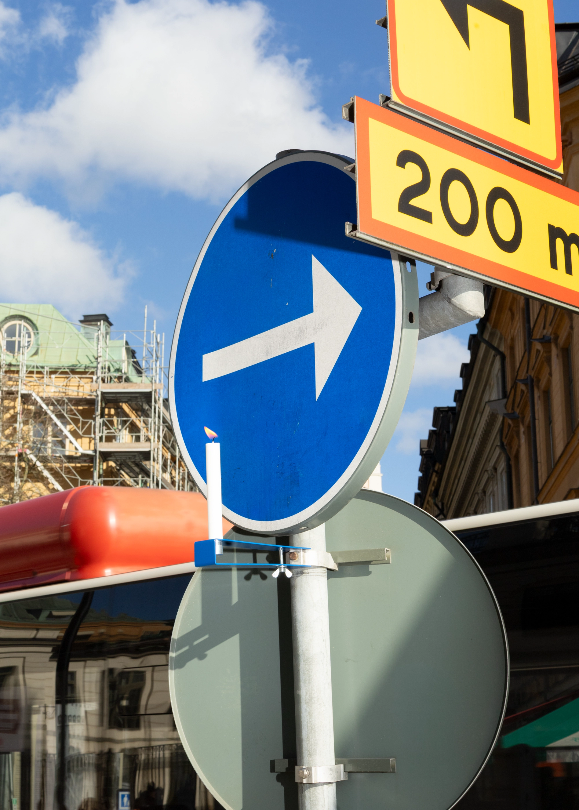 Blå ljushållare med ett vitt ljus fastklämd på en vägskyltstolpe, under en blå skylt med pil och en gul omledningsskylt.
