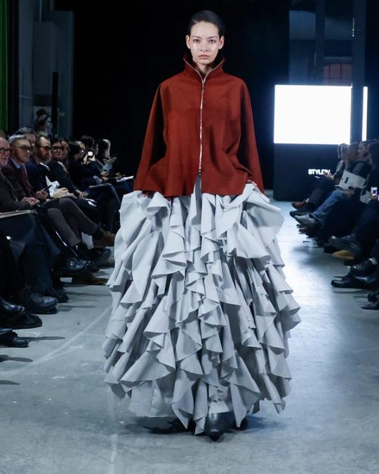 Modell på catwalk i rostbrun jacka med hög krage och en lång, voluminös grå kjol av skulpturala volanger; publik på båda sidor.