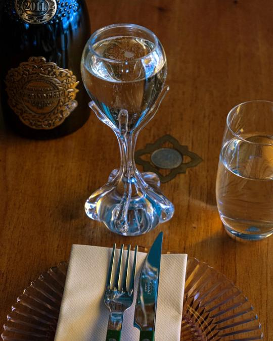 Glas på en fot av klart glas format som gripande fingrar, bredvid en flaska, ett vattenglas och bestick på en tallrik.