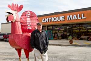 Man with big paper Mache red rooster in front of antique mall