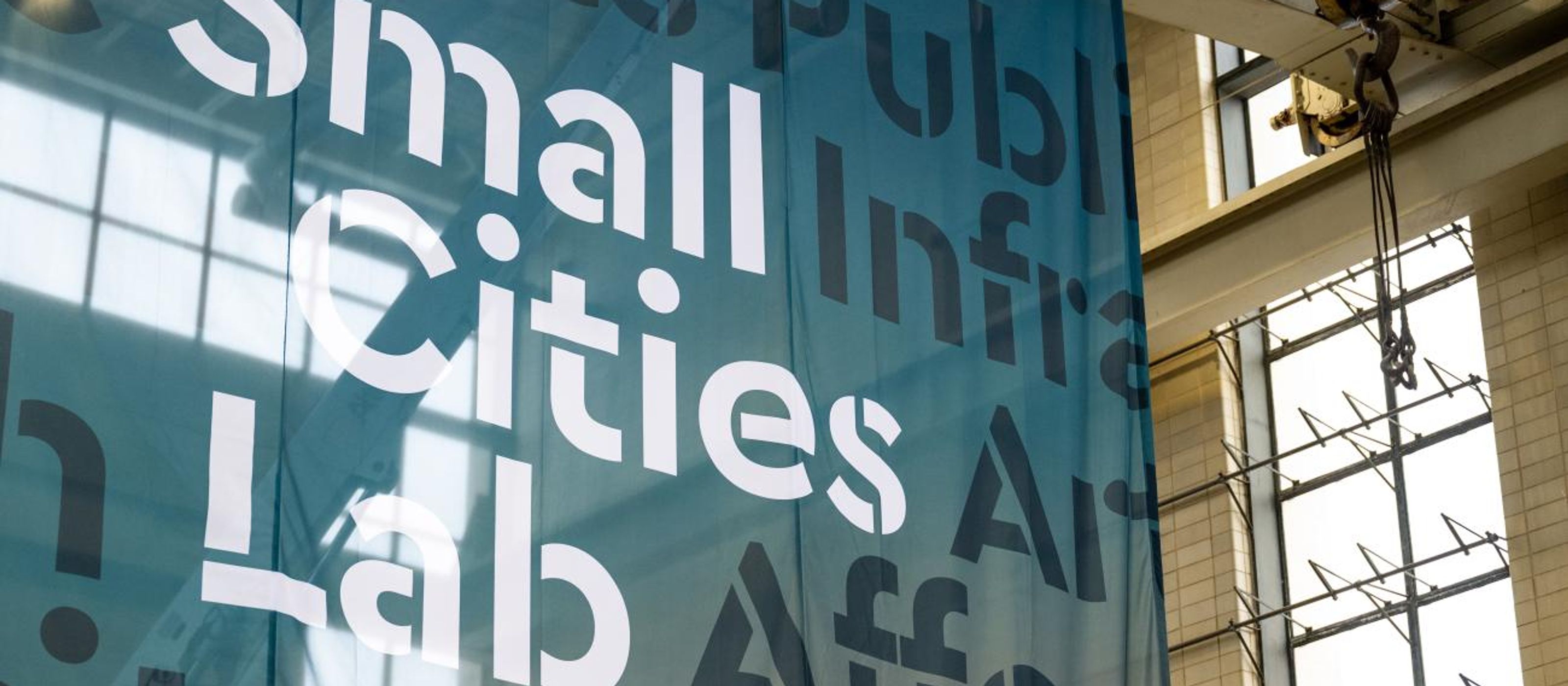 A scrim hangs in a ware house like building with "Small Cities Lab" on it