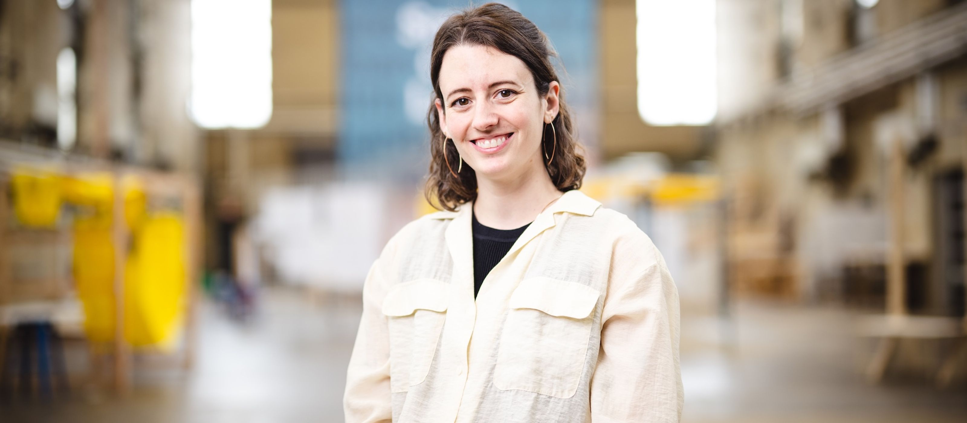 Lauren Scott stands in building C for a portrait and wears a tan colored button-up shirt.