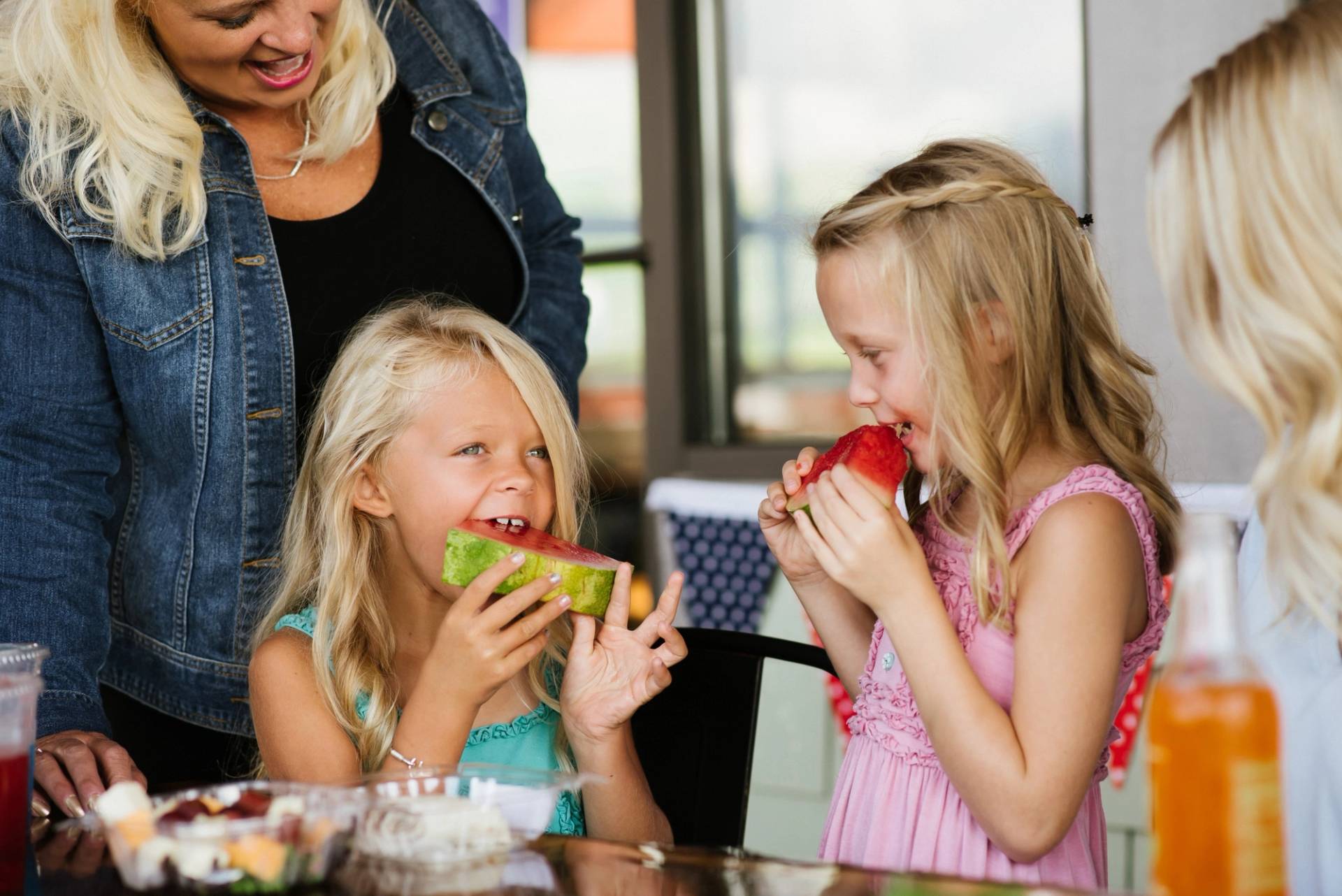 two young girls bite into slices of watermelon while their mom smiles behind them