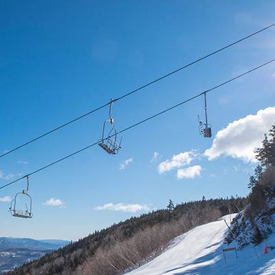 Chairs in the air from the Aurora chairlift at Sunday River, with blue skies in the background and snow on the ground.
