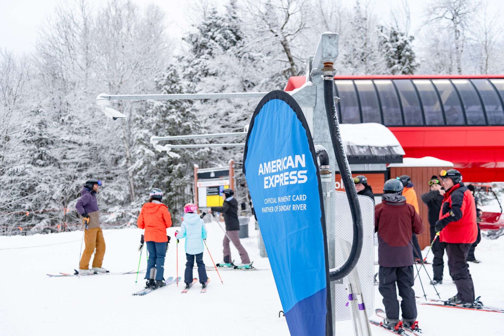 Skiers waiting in line for a chairlift at Sunday River, with an American Express flag in the ground.