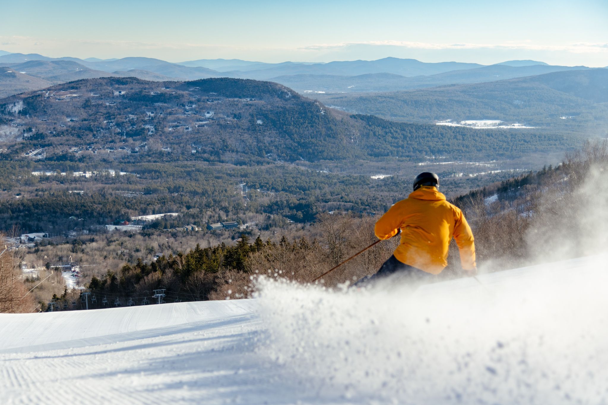 A skier in a yellow jacket skiing down a trail, with the views of the Mahoosucs, at Sunday River.