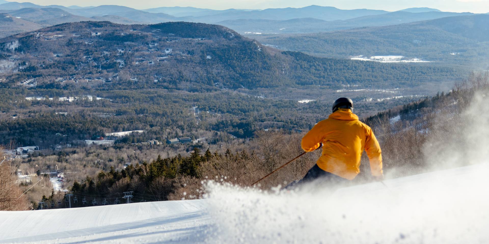 A skier in a yellow jacket skiing down a trail, with the views of the Mahoosucs, at Sunday River.