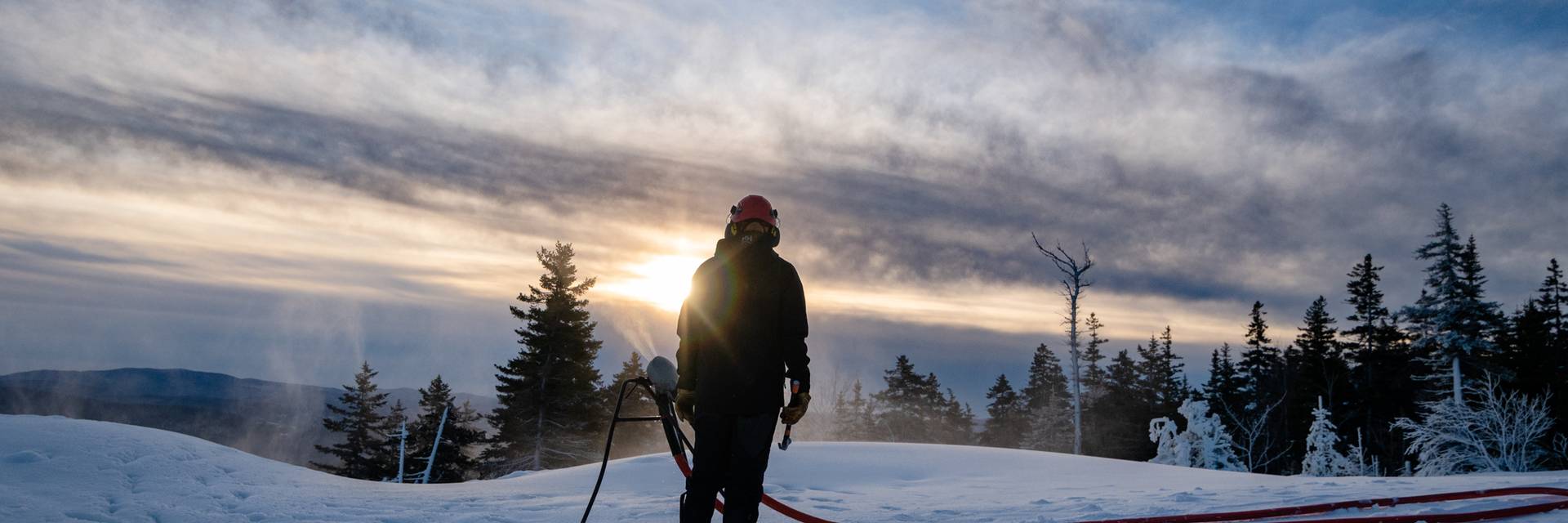 A snowmaker at Sunday River, looking at a snowgun as the sun rises.