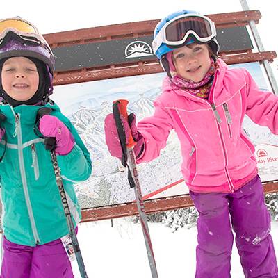 Two little girls posing in front of a sign, on skis, at Sunday River Resort.