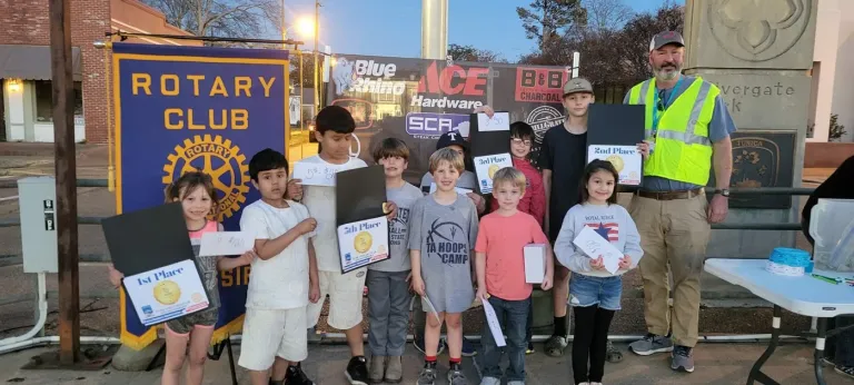 Children hold certificates beside adults and a Rotary Club banner.
