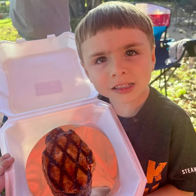 A boy holds an open foam container displaying a grilled steak.