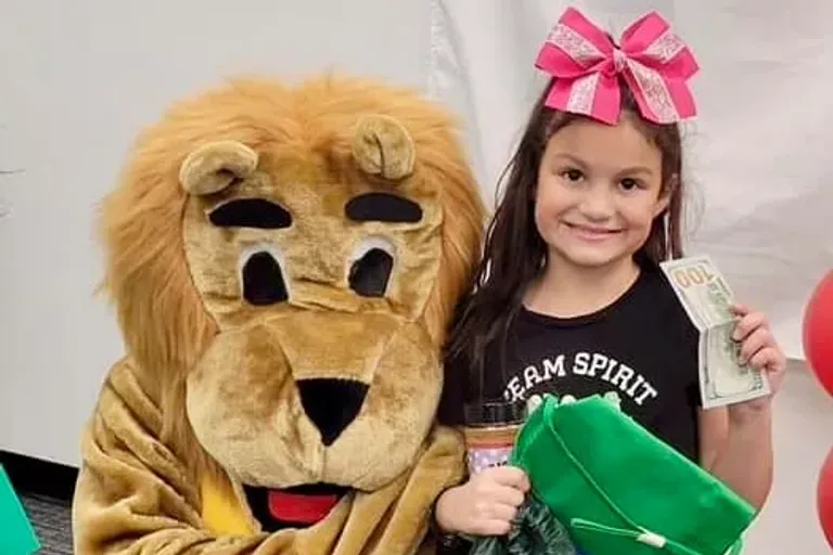 A girl with a pink bow poses beside a lion mascot while holding a prize.