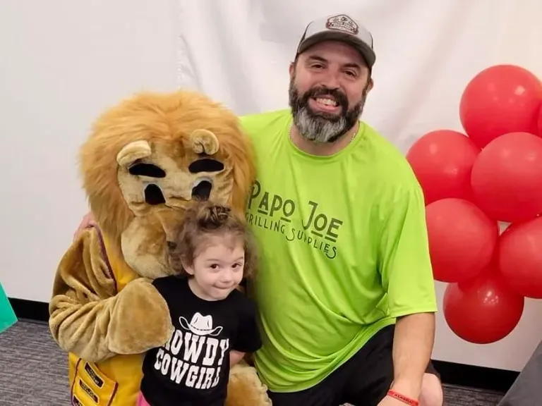 A man and a young child pose beside a lion mascot and red balloons.