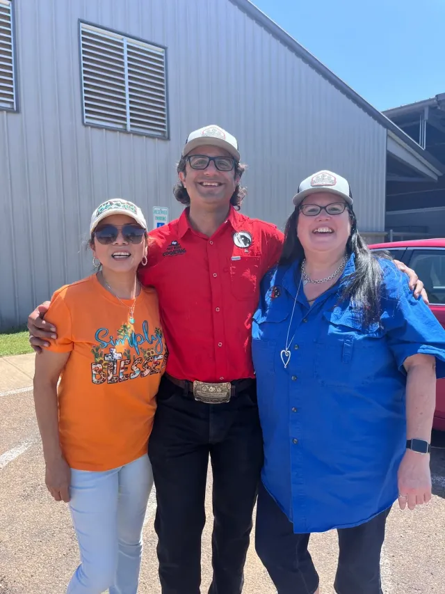 Three people in orange, red, and blue shirts pose outside a metal building.
